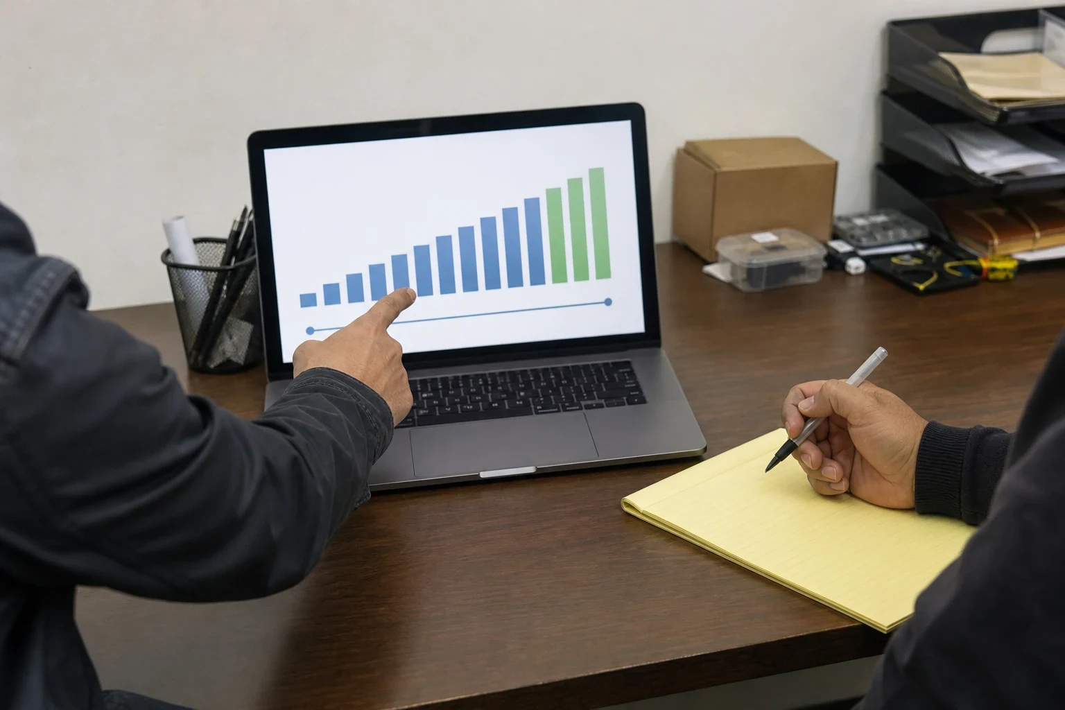 Hand pointing at a rising bar chart on a laptop at an office desk while another person holds a pen over a yellow legal pad
